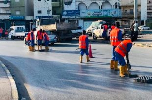 مجموعة من عمال الصيانة يرتدون سترات برتقالية وأحذية واقية أثناء صيانة وتنظيف فتحات تصريف مياه الأمطار في أحد الشوارع العامة.