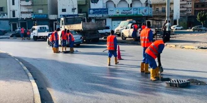 مجموعة من عمال الصيانة يرتدون سترات برتقالية وأحذية واقية أثناء صيانة وتنظيف فتحات تصريف مياه الأمطار في أحد الشوارع العامة.