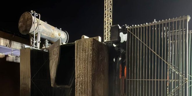 صورة ليلية لمحطة تحويل كهربائية متضررة بشدة بعد انقطاع التيار الكهربائي في المنطقة الغربية. يظهر محول كبير متفحم وسط الهياكل المعدنية، مع آثار دخان وحرارة واضحة على المكونات وأنابيب التبريد. في الخلفية، يقف برج نقل كهربائي في الظلام. الصورة توضح الأضرار المرتبطة بالإظلام الجزئي والتحقيق الجاري من قبل الشركة العامة للكهرباء.