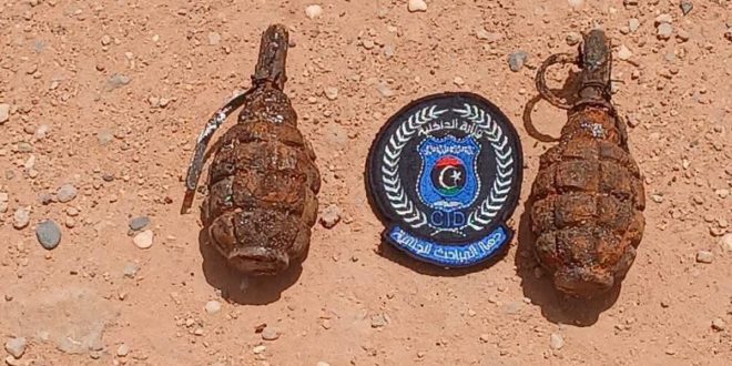 صورة لمخلفات حرب تضم قنبلتين يدويتين صدئتين وبينهما شعار جهاز المباحث الجنائية على أرض ترملية في سبها.
