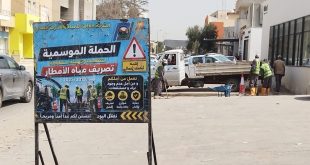صورة خارجية نهارية توضح أعمال تنظيف خزان تجميع مياه الأمطار بمنطقة السبعة، حيث تقف فرق الشركة العامة للمياه والصرف الصحي (GCMWS) خلف لافتة كبيرة للحملة الموسمية. العمال يرتدون سترات عاكسة ويقفون بجوار شاحنات الخدمات وصهريج شفط مياه الصرف الصحي على جانب الشارع. المباني السكنية والتجارية واضحة في الخلفية.