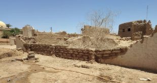 لقطة واسعة لبقايا جدران ومبانٍ أثرية مبنية من الطوب اللبن والطين تحت أشعة الشمس في مدينة غات، وتظهر في الخلفية قبة مسجد وأشجار نخيل.