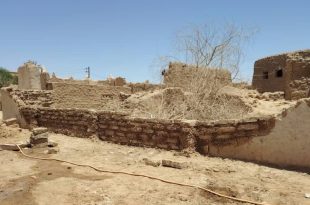 لقطة واسعة لبقايا جدران ومبانٍ أثرية مبنية من الطوب اللبن والطين تحت أشعة الشمس في مدينة غات، وتظهر في الخلفية قبة مسجد وأشجار نخيل.