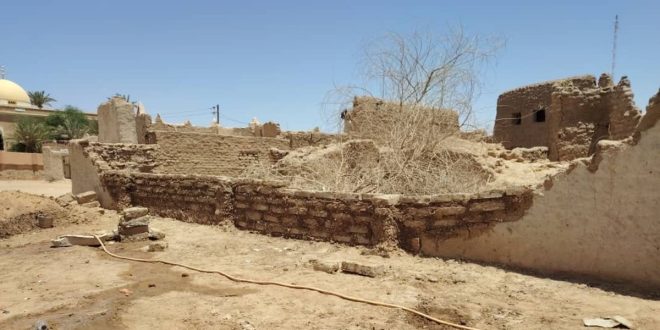 لقطة واسعة لبقايا جدران ومبانٍ أثرية مبنية من الطوب اللبن والطين تحت أشعة الشمس في مدينة غات، وتظهر في الخلفية قبة مسجد وأشجار نخيل.