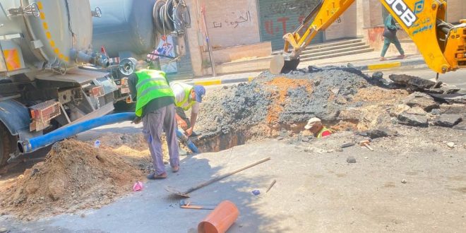 عمال وصيانة يرتدون سترة فسفورية يمارسون أعمال حفر وتغيير أنبوب داخل حفرة في الطريق العام بشارع الصريم،