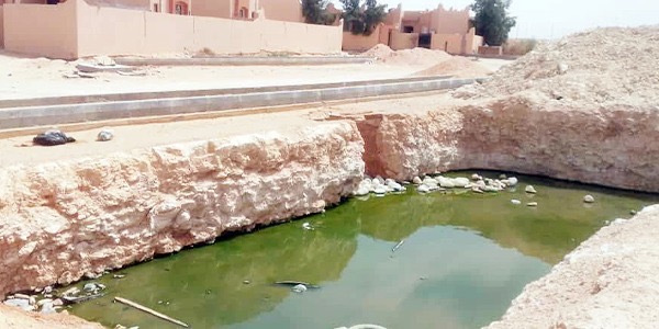 حفرة مكشوفة كبيرة في منطقة ترابية أمام مبانٍ سكنية، تتجمع فيها مياه الصرف الصحي ذات اللون الأخضر والداكن.