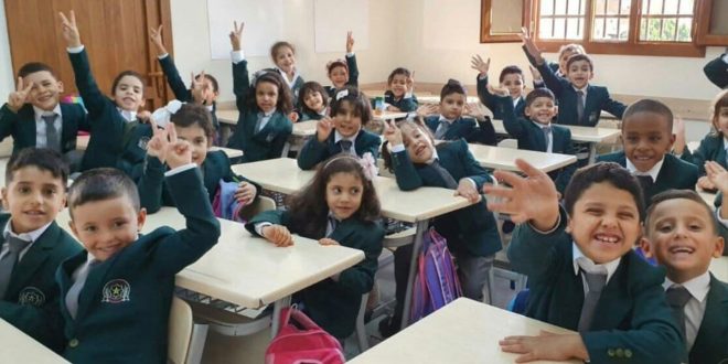 مجموعة من الأطفال والطلاب الليبيين يجلسون داخل الفصل الدراسي مرتديين الزي المهرجاني/المدرسي ويوجهون علامات النصر والترحيب للكاميرا.