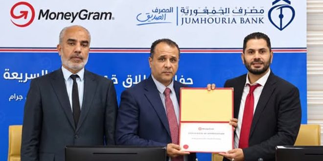 المدير العام المكلف لمصرف الجمهورية ومسؤولون آخرون يقفون جنبًا إلى جنب أثناء توقيع أو التقاط صورة تذكارية مع وثيقة اتفاقية الشراكة مع موني غرام أمام خلفية تحمل شعاري مصرف الجمهورية وMoneyGram.