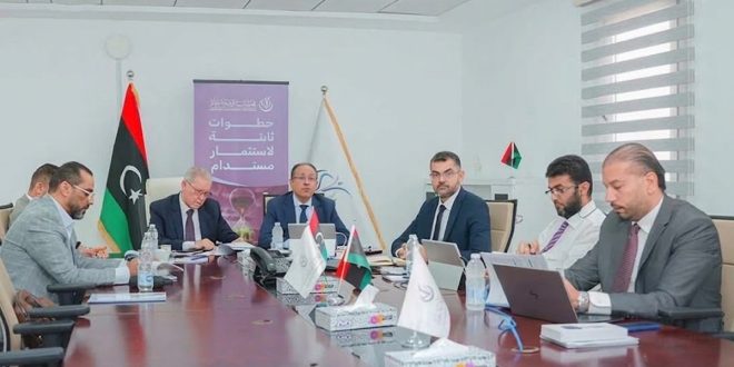 صورة لاجتماع رسميين ومسؤولين يجلسون حول طاولة اجتماعات بيضاوية في قاعة مؤتمرات، وأمامهم أجهزة حاسوب محمولة وأوراق، وفي الخلفية يظهر علم ليبيا إلى جانب لافتة مدون عليها عبارة "خطوات ثابتة لاستثمار مستدام".