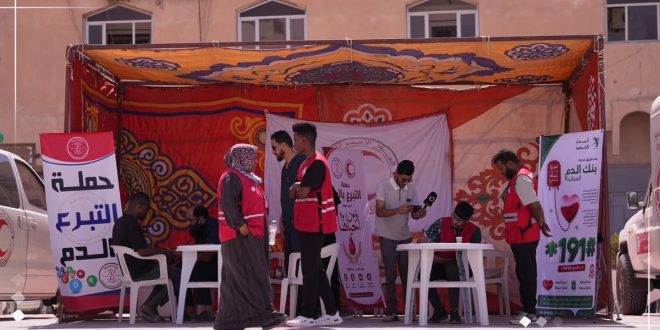 متطوعون يرتدون سترات الهلال الأحمر الليبي الأحمر يقفون أمام خيمة حملة تبرع بالدم مزينة بالأعلام، بينما يسجل شخص آخر بياناته على طاولة في المقدمة، وعلى اليسار لوحة إعلانية كبيرة مكتوب عليها باللغة العربية "حملة التبرع بالدم".