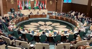 لقطة زاوية مرتفعة واسعة لقاعة اجتماعات جامعة الدول العربية، يظهر فيها الوفود العربية يجلسون حول طاولة دائرية كبيرة تحيط بها أعلام الدول العربية