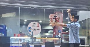 عنصر من جهاز الحرس البلدي يرتدي زيه الرسمي الأزرق والداكن وقبعة، ينزل الستار المعدني لمحل أو مطعم مغلق من واجهته الزجاجية، وتظهر على الزجاج ملصقات لإعلانات خدمات الدفع الإلكتروني وخدمات التوصيل