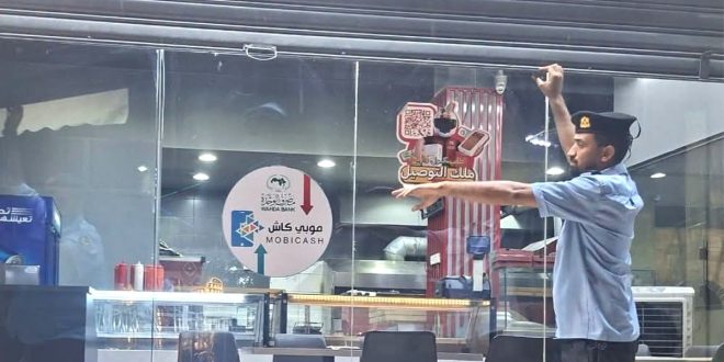 عنصر من جهاز الحرس البلدي يرتدي زيه الرسمي الأزرق والداكن وقبعة، ينزل الستار المعدني لمحل أو مطعم مغلق من واجهته الزجاجية، وتظهر على الزجاج ملصقات لإعلانات خدمات الدفع الإلكتروني وخدمات التوصيل