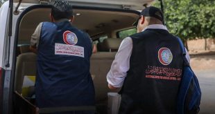 شخصان من الكوادر الطبية يرتديان سترة كحيلة يحملان شعار المركز الوطني لمكافحة الأمراض أثناء إعداد معداتهما بالقرب من سيارة نقل ميدانية.