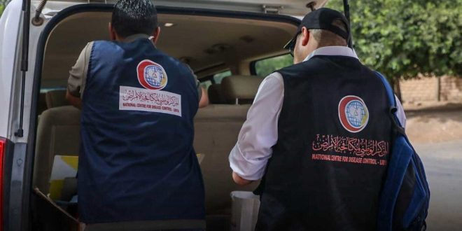 شخصان من الكوادر الطبية يرتديان سترة كحيلة يحملان شعار المركز الوطني لمكافحة الأمراض أثناء إعداد معداتهما بالقرب من سيارة نقل ميدانية.