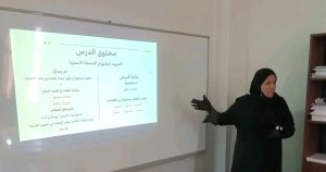 مترشحة لمهنة التفتيش التربوي تشرح عرضاً تقديمياً حول محتوى درس عن الجملة الاسمية باستخدام البروجكتر أمام لجنة التقييم.