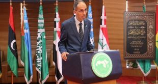 متوسط العمر بنظارات وبدلة رسمية كحلية يلقي كلمة خلف منبر ينشر شعار المنظمة العربية للتربية والثقافة والعلوم (الألكسو). تحيط به أعلام دول عربية، وإلى جانبه لوحة تكريمية مؤطرة تحمل غلاف كتابه المحقق "الغيث الذي انسجم في شرح لامية العجم