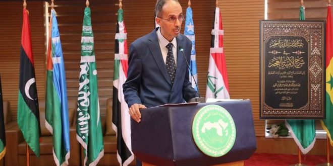 متوسط العمر بنظارات وبدلة رسمية كحلية يلقي كلمة خلف منبر ينشر شعار المنظمة العربية للتربية والثقافة والعلوم (الألكسو). تحيط به أعلام دول عربية، وإلى جانبه لوحة تكريمية مؤطرة تحمل غلاف كتابه المحقق "الغيث الذي انسجم في شرح لامية العجم