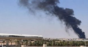 لقطة خارجية نهارية تُظهر عموداً هائلاً من الدخان الأسود الداكن يتصاعد في السماء فوق منطقة منشآت حيوية، مع ظهور جزء من محطة قطار المطار على اليسار وبعض النخيل في المقدمة