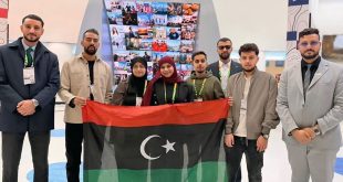 وفد شبابي ليبي رسمي يضم ثمانية أفراد يرفعون علم ليبيا في مهرجان الشباب الدولي بروسيا. جميعهم يرتدون بطاقات تعريف ويقفون أمام شاشة عرض كبيرة في قاعة مؤتمرات حديثة.