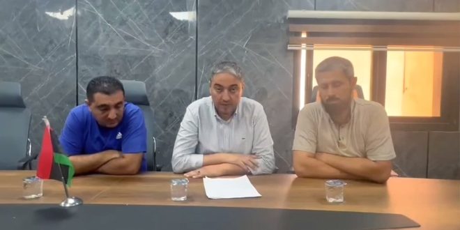 ثلاثة مسؤولين من إدارة مستشفى يفرن يجلسون خلف طاولة اجتماعات خشبية يضعون عليها ورقة بيان رسمي وعلم ليبيا أثناء إلقاء بيان مصور.