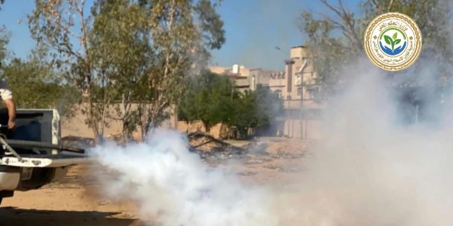 سيارة رش ورذاذ الدخان الأبيض يغطي المكان أثناء عمليات التعقيم والرش لمكافحة الآفات والحمى القلاعية في جنوب طرابلس وشعار مديرية شؤون الإصحاح البيئي يظهر في الأعلى.