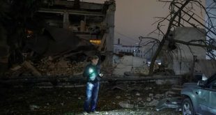 رجل يقف ليلاً وسط حطام مبنى مدمر ناتج عن قصف جوي ويضيء المكان باستخدام كشاف ضوئي محمول