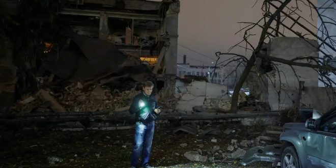 رجل يقف ليلاً وسط حطام مبنى مدمر ناتج عن قصف جوي ويضيء المكان باستخدام كشاف ضوئي محمول