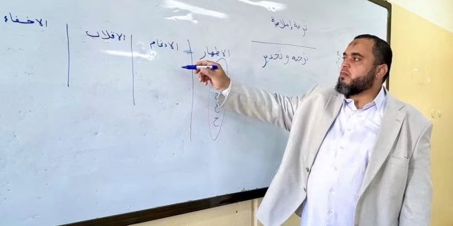 معلم أو مسؤول تربوي يقف أمام سبورة بيضاء ويكتب عليها بقلم العرض خلال متابعة سير الدراسة في إحدى المدارس.