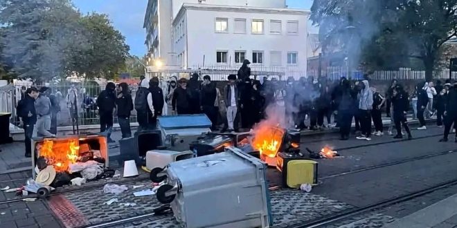 طلاب غاضبون ومقنعون يقفون في الشارع خلف حاجز من حاويات القمامة المشتعلة والدخان يتصاعد منها أثناء الاحتجاجات المدرسية في فرنسا.