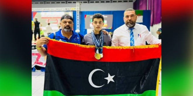 الرباع الليبي عبدالرحيم عزالدين صالح يرتدي الميداليات ويتوسط شخصين يمسكون بالعلم الليبي خلال تتويجه في بطولتي أفريقيا والعرب لرفع الأثقال بمدينة مستغانم الجزائرية