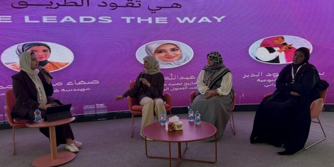 ​لقطة عرضية لمنصة حوارية تجلس عليها أربع سيدات ليبيات محجبات يشاركن في ندوة. خلفهن شاشة عرض أرجوانية كبيرة مضاءة مكتوب عليها باللغتين العربية والإنجليزية: "هي تقود الطريق" و "SHE LEADS THE WAY". تتحدث السيدة التي على اليسار في الميكروفون، بينما تستمع إليها الثلاث الأخريات الجالسات على كراسي وردية مريحة. أمامهن طاولات قهوة صغيرة عليها زجاجات مياه ومحارم. المشهد يمثل إطلاق مبادرة "رايس" للتمكين الرقمي في طرابلس.