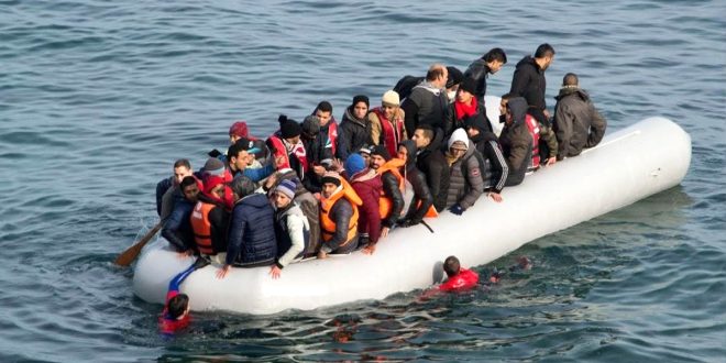 ​صورة ملتقطة من زاوية علوية تُظهر قاربًا مطاطيًا أبيض ومكتظًا بأكثر من 20 مهاجرًا في عرض البحر المتوسط. يرتدي العديد منهم سترات نجاة برتقالية، وتُظهر الصورة أيضًا شخصين (منقذين أو مهاجرين) يسبحان في الماء بالقرب من القارب.