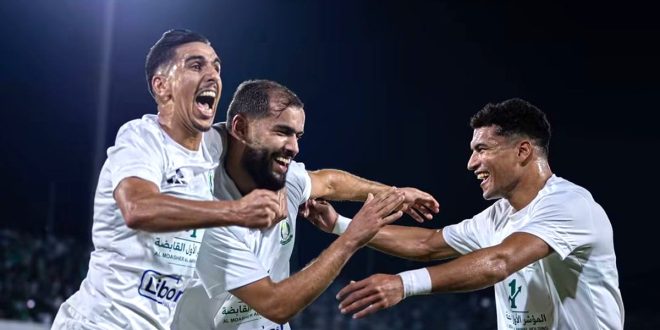 ​ثلاثة لاعبين من فريق الأهلي طرابلس بملابس رياضية بيضاء يحتفلون بفرح شديد بعد تسجيل هدف في مباراة تحت الأضواء الكاشفة في ملعب طرابلس الدولي ضد فريق ويلوالو الإثيوبي في كأس الكونفدرالية الإفريقية.