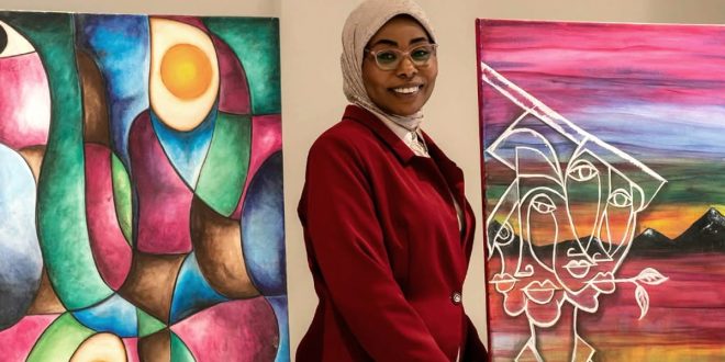​الفنانة التشكيلية والفوتوغرافية الليبية هند الراشدي تقف مبتسمة بين لوحتين من أعمالها الفنية التشكيلية ذات الألوان الزاهية والأشكال التجريدية، خلال مشاركتها في معرض فني يدمج بين التصوير الفوتوغرافي والفن التشكيلي