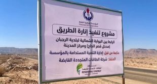 ​صورة للوحة إعلانية كبيرة في موقع مشروع، تُعلن عن قيام المؤسسة الوطنية للنفط بتسليم موقع مشروع لإنارة الطريق بالطاقة الشمسية بين قصر الحاج والرجبان. تظهر اللوحة تفاصيل المشروع، بما في ذلك شعارات المؤسسة وشركة الطاقات المتجددة المنفذة، وموقع المشروع، في بيئة صحراوية مفتوحة بجانب طريق معبد.