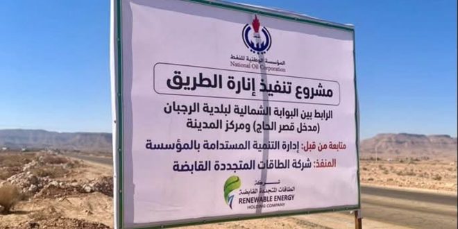 ​صورة للوحة إعلانية كبيرة في موقع مشروع، تُعلن عن قيام المؤسسة الوطنية للنفط بتسليم موقع مشروع لإنارة الطريق بالطاقة الشمسية بين قصر الحاج والرجبان. تظهر اللوحة تفاصيل المشروع، بما في ذلك شعارات المؤسسة وشركة الطاقات المتجددة المنفذة، وموقع المشروع، في بيئة صحراوية مفتوحة بجانب طريق معبد.