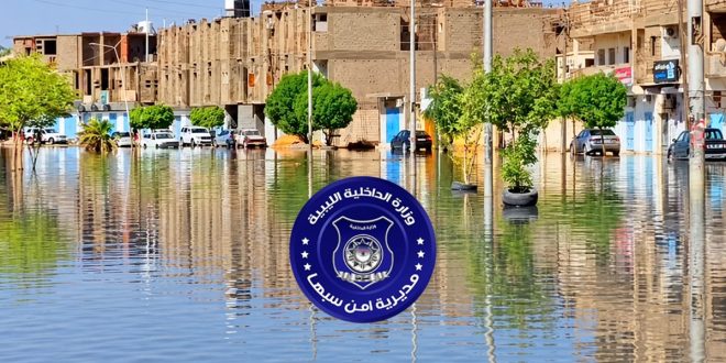 صورة فوتوغرافية لتجمعات مياه الأمطار تغمر شوارع مدينة سبها مع شعار مديرية أمن سبها في المقدمة
